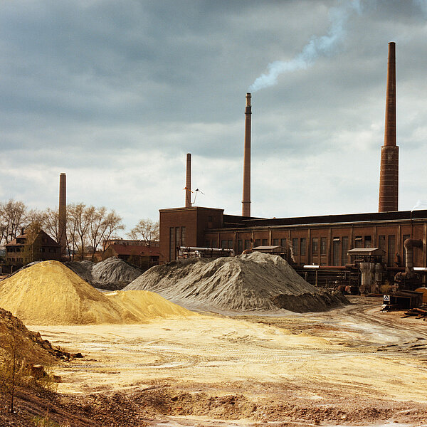 Fabrik mit rauchenden Schornsteinen und sandigem Boden im Vordergrund