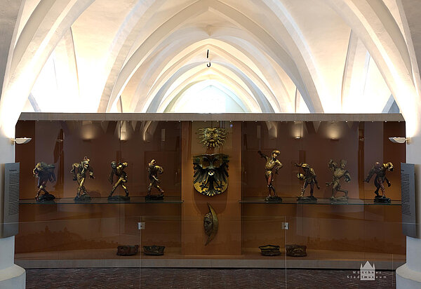 Glasvitrine mit acht Holzfiguren sowie drei geschnitzten Wappen.