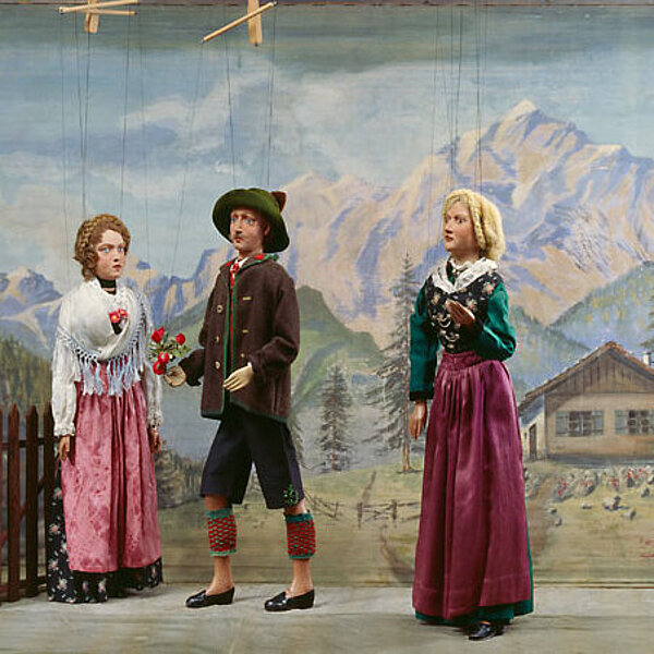 Vier Marionetten in traditionell bayerischer Kleidung, zwei Männer und zwei Frauen vor alpinem Hintergrund.