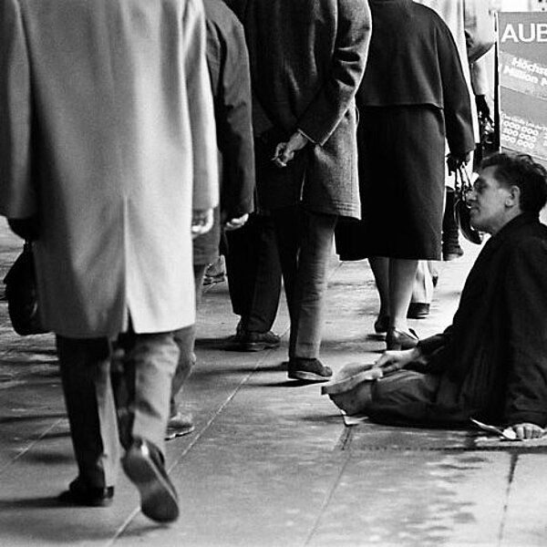 Schwarzweiß-Fotografie eines bettelnden Mannes, der neben den vorbeilaufenden Passanten am Boden sitzt