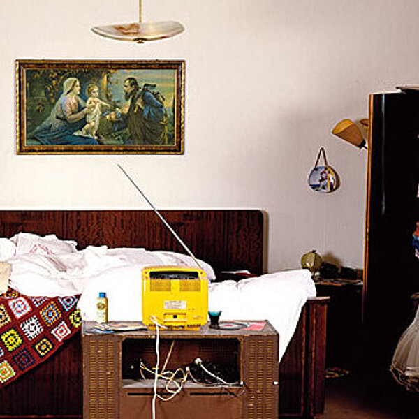 Ansicht eines Schlafzimmers im Stil der 70er Jahre mit zerwühltem Bett und herumliegender Kleidung.