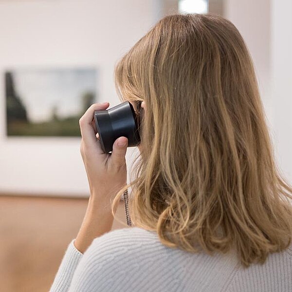 Eine junge Frau in Rückenansicht hält sich in einer Ausstellung einen Einhandhörer ans Ohr.