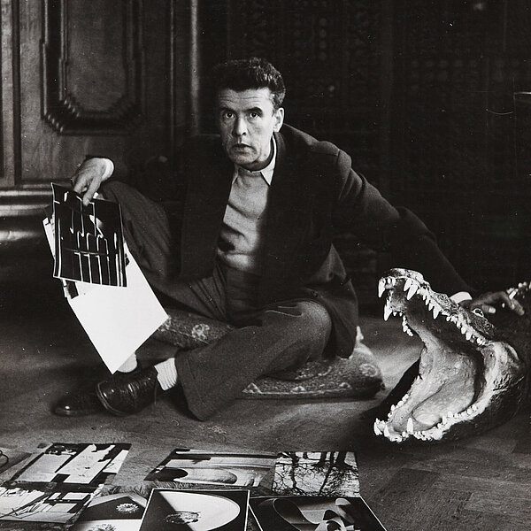 Ein Mann sitzt inmitten von Fotografien auf dem Boden neben einem Krokodil mit aufgerissenem Maul
