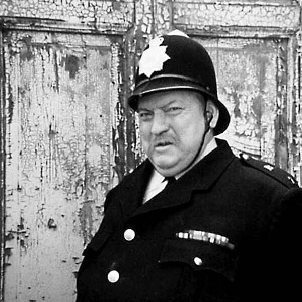 Ein englischer Polizist steht vor einer abblätternden Fassade und blickt grimmig in die Kamera.