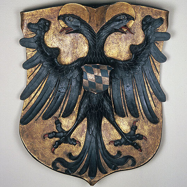 Holzwappen mit doppelköpfigen Vogel, der ein blau-weiß kariertes Wappen auf der Brust trägt, Vogel in Schwarz, Wappenhintergrund in Gold bemalt.