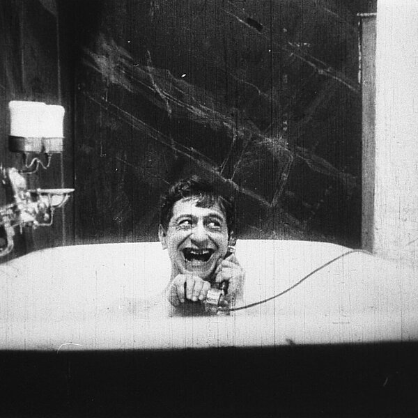 Ein junger Mann sitzt grinsend in einer Badewanne und telefoniert.