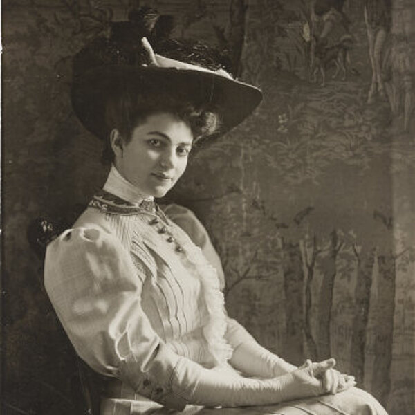 Junge Frau mit Bluse, langem Rock, weißen Handschuhen und Hut, vor einer Bildtapete im Fotostudio sitzend