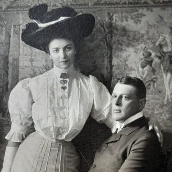 Stehende junge Frau mit Hut blickt den Betrachter an und legt ihrem sitzenden Ehemann die Hand auf die Schulter.