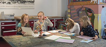Vier Mädchen im Grundschulalter sitzen an einem Tisch, auf dem Lachende Kinder sitzen an einem Tisch und zeichnen.Holzfigur eines Moriskentänzers in gold-rotem Gewand steht.